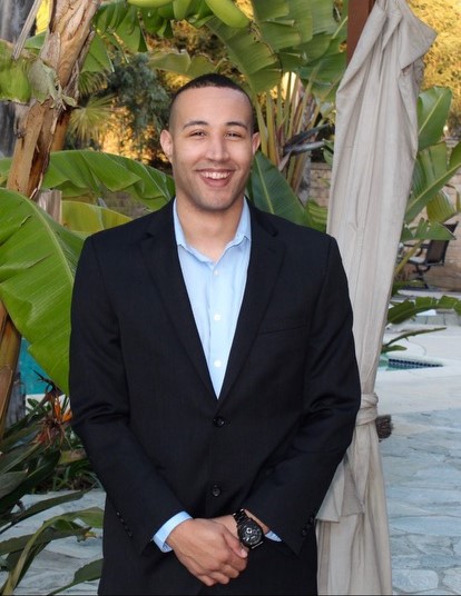 Cameron McFadden - Man wearing a dark suit jacket and light blue, collared dress shirt, standing and smiling, facing the camera.